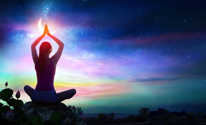 A young woman sits in a yoga pose facing away from the viewer. She is looking into a beautiful evening sky with a crescent moon and many colors at the horizon.